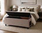 A beige upholstered storage bench with a lift-up lid, shown in a styled bedroom setting in front of a bed with an opening in the side for a pet entrance.