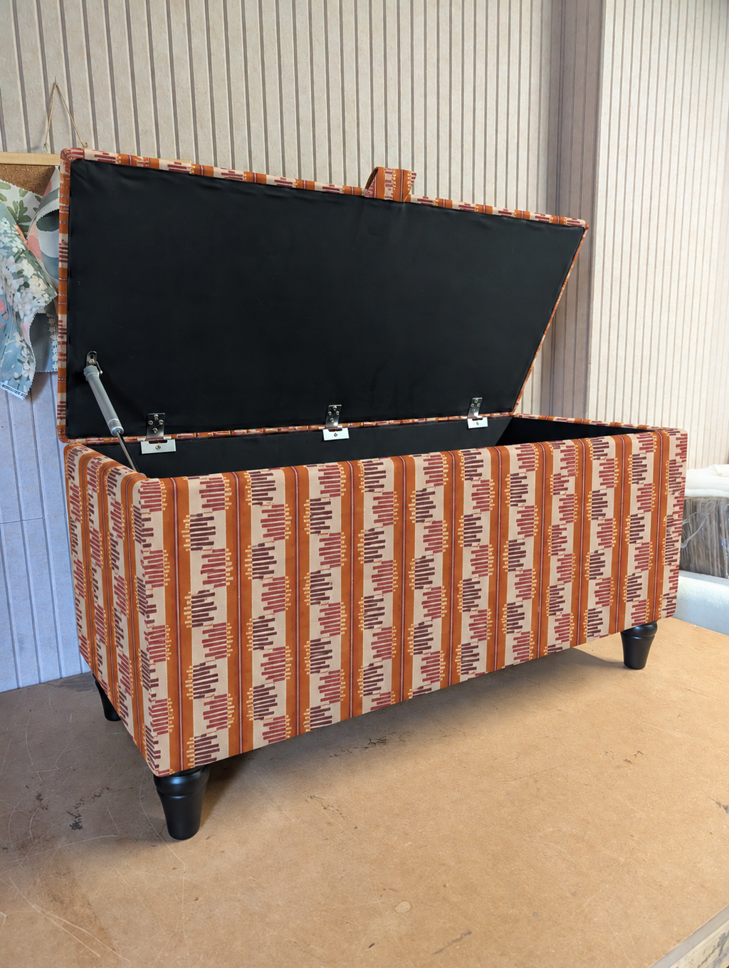 An upholstered storage bench with a hinged lid, featuring a rust-orange, beige, and brown geometric patterned fabric. The bench is shown open, revealing a black fabric-lined interior with metal gas lift supports.