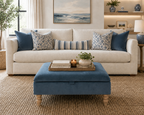 A styled living room scene featuring a cream-colored upholstered sofa with blue and patterned throw pillows, a matching blue ottoman coffee table in the foreground, and a woven jute rug on the floor.