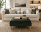 A styled living room scene featuring a cream-colored upholstered sofa with multiple decorative throw pillows, a dark green velvet ottoman in the foreground, and a neutral-toned woven rug on the floor.