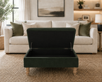 A dark forest green upholstered storage ottoman with a lift-up lid and turned wooden legs, shown in a bright, modern living room in front of a cream sofa.