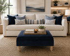 A styled living room scene featuring a cream-colored sofa with navy and patterned throw pillows, a matching navy ottoman coffee table in the foreground, and a neutral-toned interior with wall art and shelving.