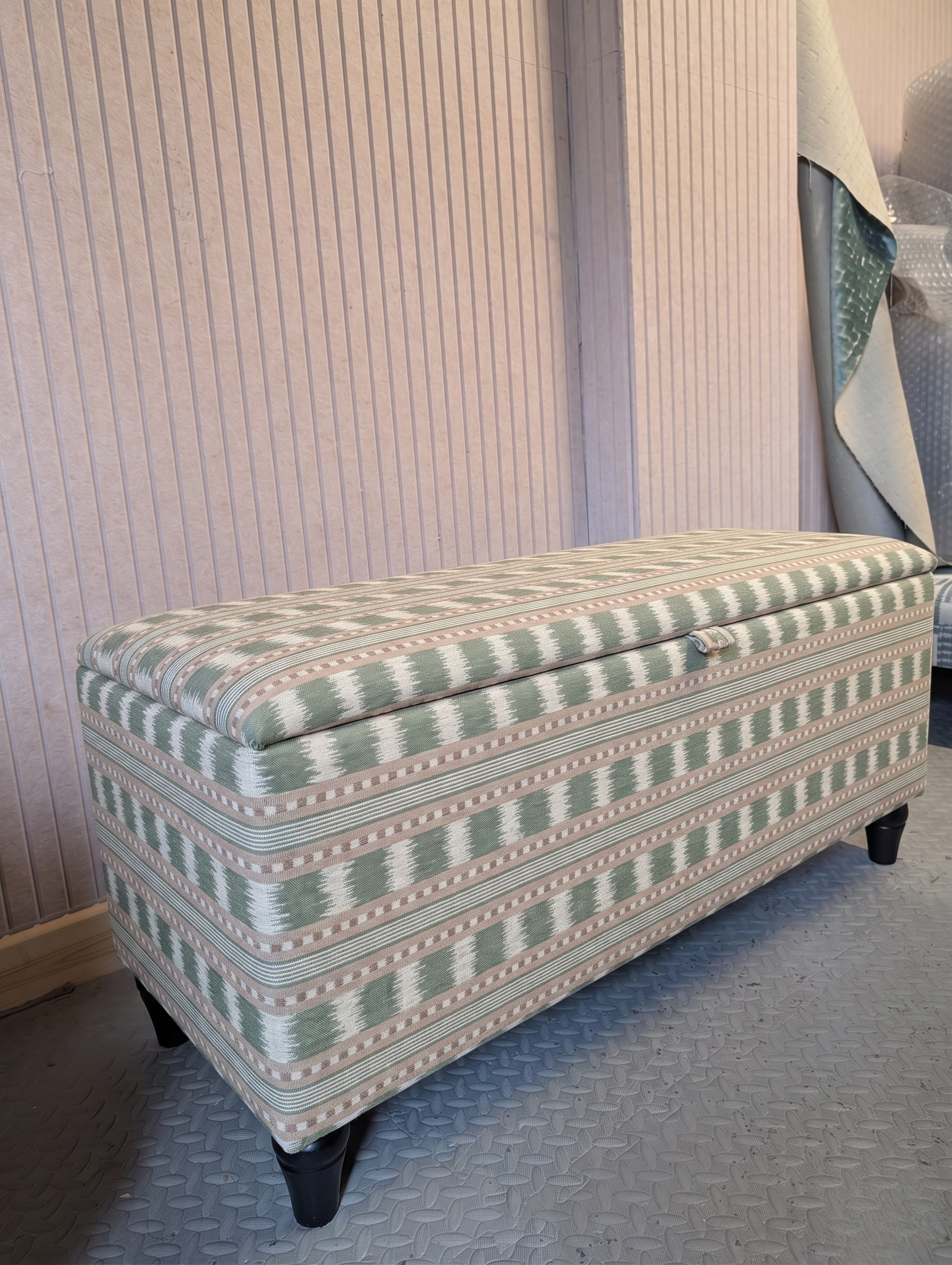 An upholstered storage ottoman in James hare Paxton fabric green and pink colourway. Stay in a workshop room with Grey textured floor and pink textured walls.