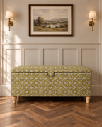 A storage bench with a green and beige floral patterned fabric upholstery, styled in a traditional room setting with a wooden herringbone floor, white wainscoting, and wall sconces.