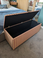 A striped fabric storage bench with its lid propped open, showing a black interior. The bench has vertical stripes in dusty pink and a yellowcolor, set on a carpeted floor in a bedroom next to a bed.