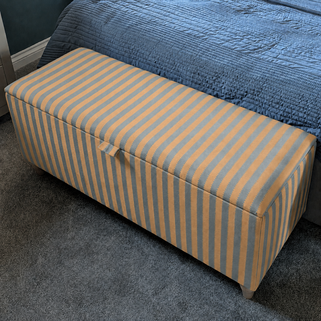 A rectangular upholstered storage bench with vertical mustard yellow and blue stripes, positioned at the foot of a bed with a blue quilted cover in a bedroom setting.