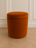 A round, barrel-shaped upholstered ottoman stool in a rich burnt orange velvet fabric, set against a neutral cream paneled wall and light wood herringbone floor.