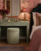 Round sage green upholstered storage dressing stool with a padded lift up lid, positioned beneath a matching sage green dressing table. The bedroom is decorated with pink floral wallpaper, a scalloped gold mirror, brass table lamp, dark green upholstered bed and layered blush pink bedding.
