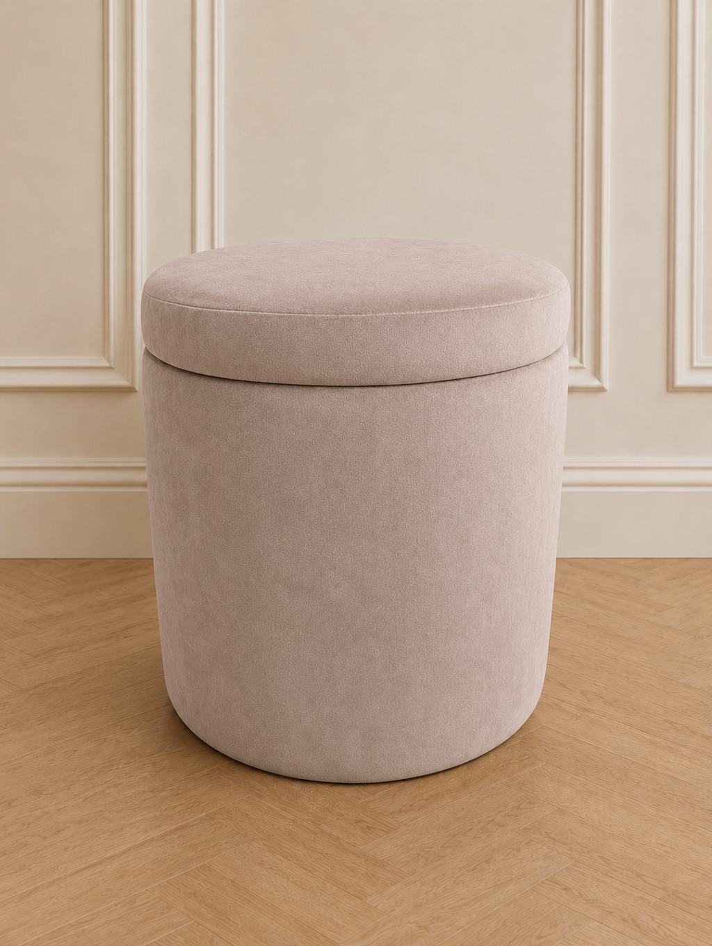 A cylindrical upholstered storage ottoman stool in a light beige fabric, set against a cream paneled wall and light wood herringbone floor.