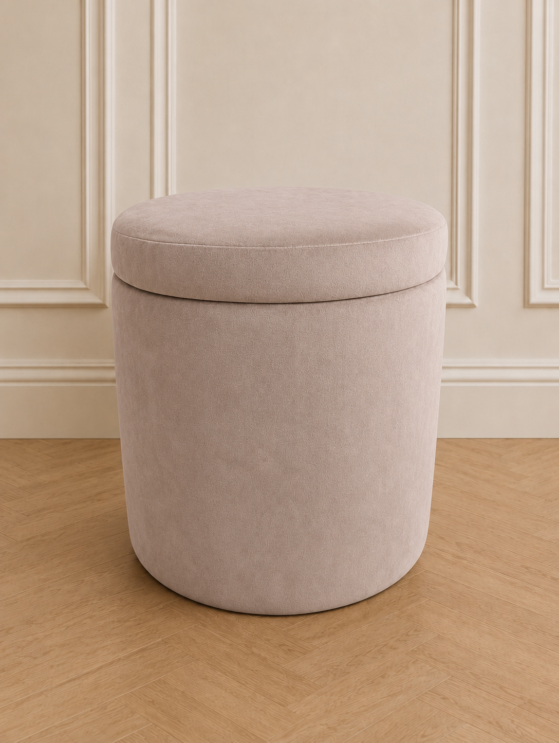 A cylindrical upholstered storage ottoman stool in a light beige fabric, set against a cream paneled wall and light wood herringbone floor.