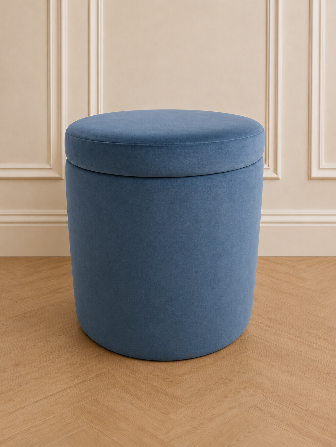 A round upholstered storage ottoman stool in a dusty medium blue velvet fabric, set against a cream paneled wall and light wood herringbone floor.