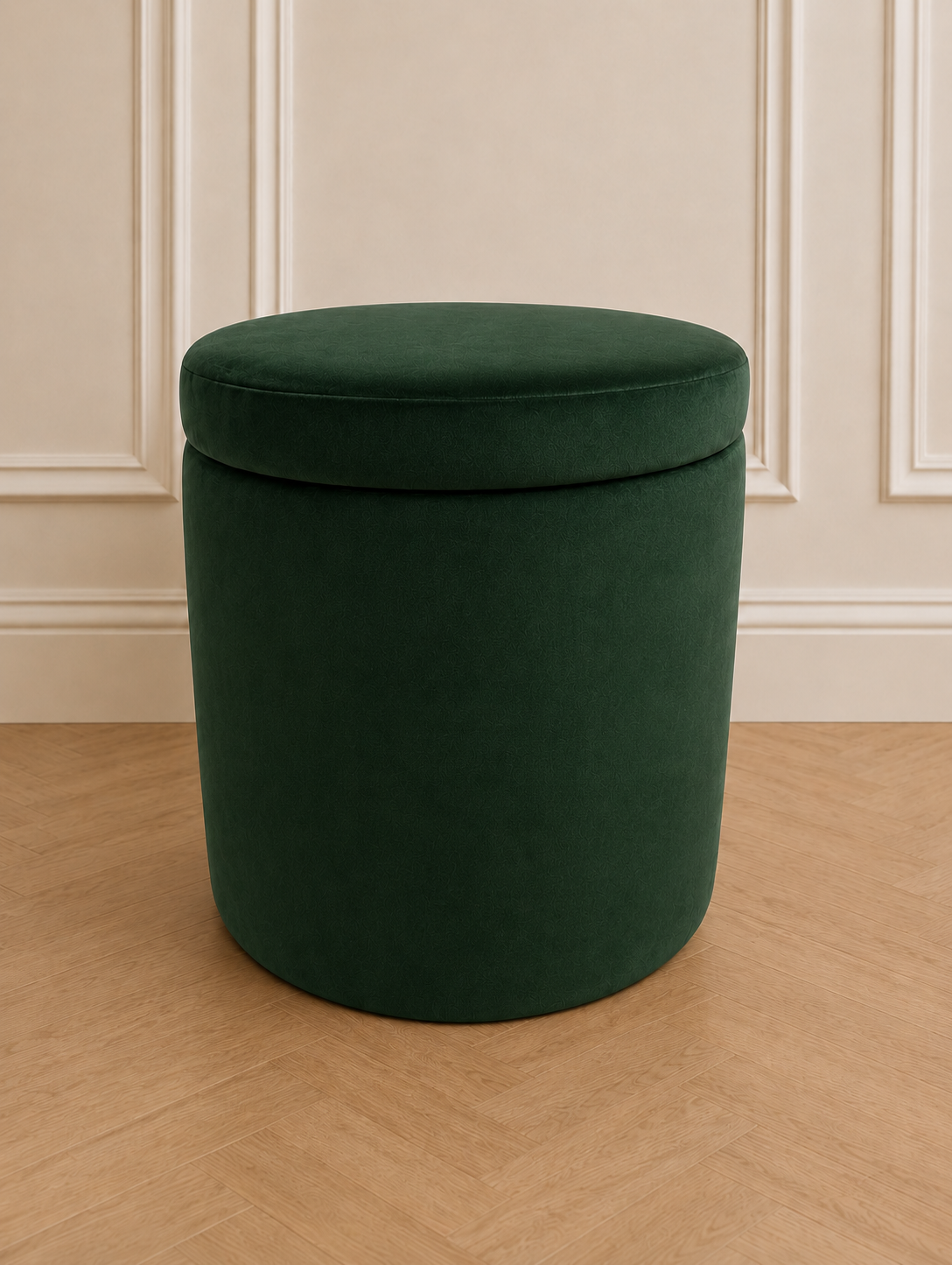 A round, upholstered storage ottoman in deep emerald green velvet, set against a cream paneled wall and light wood herringbone floor.