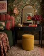 A styled bedroom interior featuring a black console table with a gold-framed mirror, a mustard yellow cylindrical ottoman, green floral wallpaper, and a pink upholstered bed with patterned pillows.