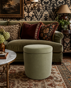 A styled interior scene featuring an olive green velvet cylindrical ottoman in the foreground, with a green sofa, patterned throw pillows, floral wallpaper, a rug, and side tables in the background.
