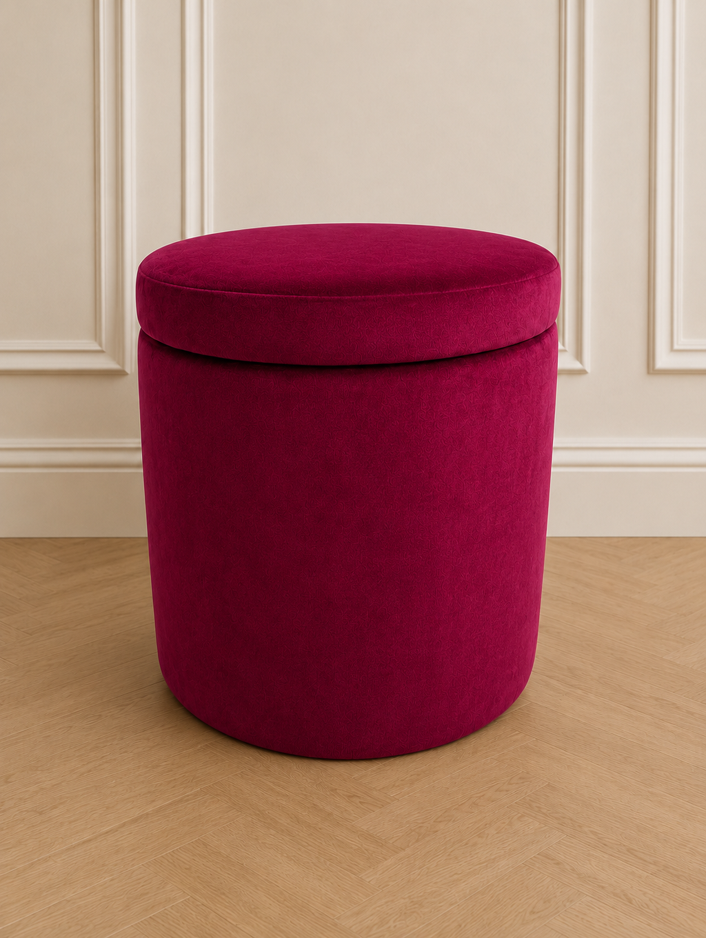 A cylindrical upholstered ottoman stool in rich magenta velvet, shown on a light wood floor against a cream paneled wall.