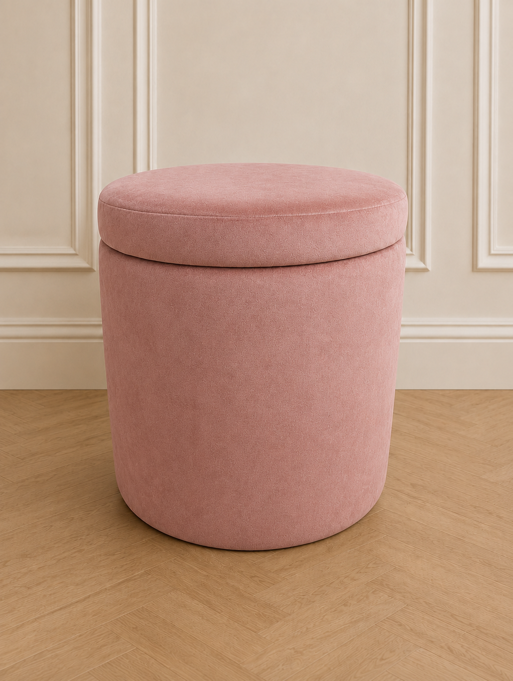 A round upholstered storage ottoman stool in rose pink velvet, shown on a light wood herringbone floor against a cream panelled wall.