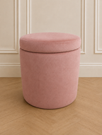 A round upholstered storage ottoman stool in rose pink velvet, shown on a light wood herringbone floor against a cream panelled wall.