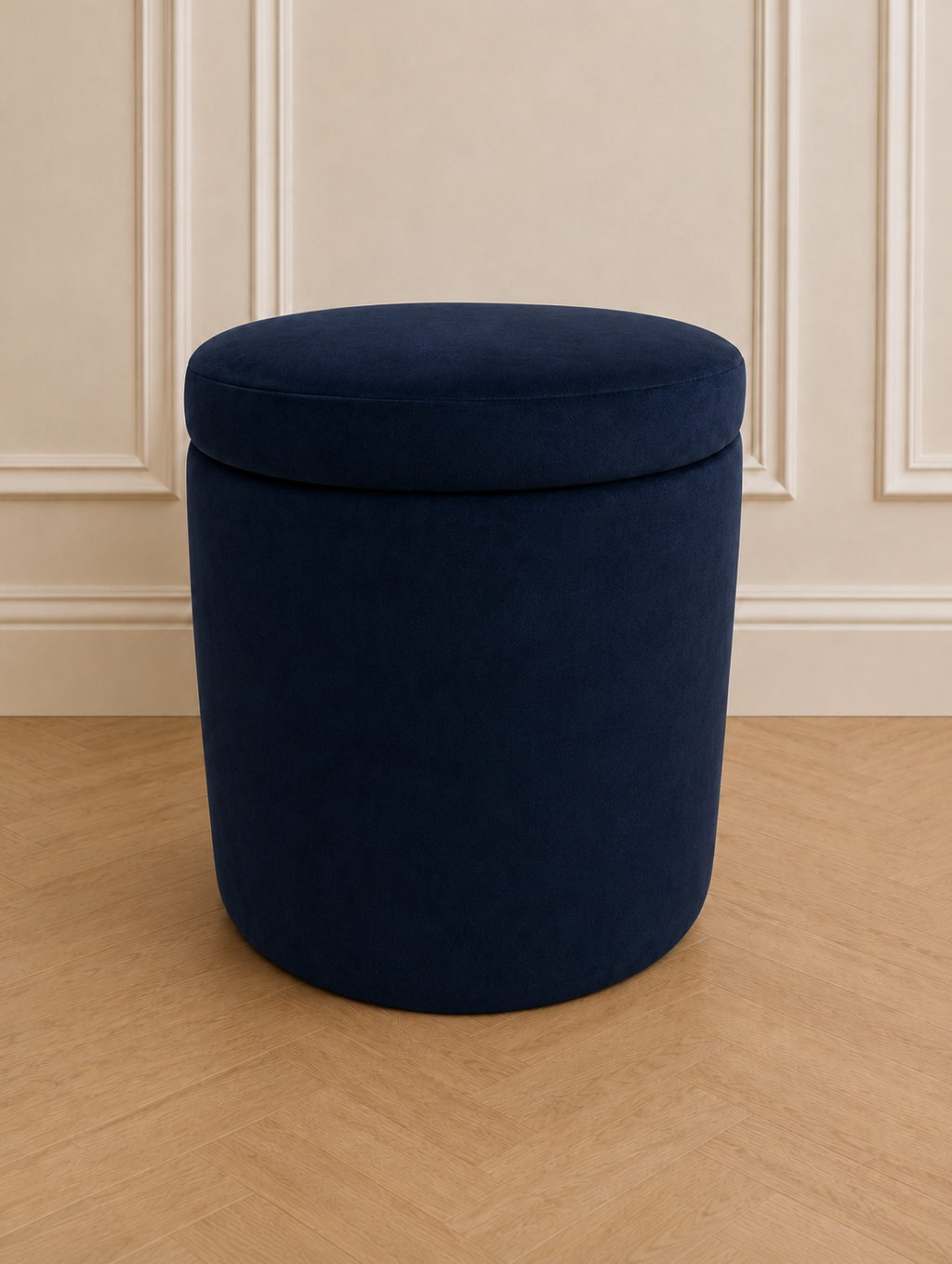 A round upholstered ottoman stool in deep Royal blue velvet, shown in a room setting with a light wood herringbone floor and cream paneled wall.