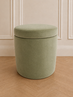 A round, upholstered storage ottoman in a muted sage green velvet fabric, set against a cream paneled wall and light wood herringbone floor.