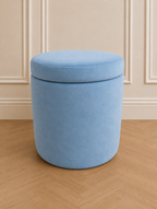 A light blue velvet cylindrical storage ottoman with a cushioned lid, set on a light wood herringbone floor against a beige paneled wall.