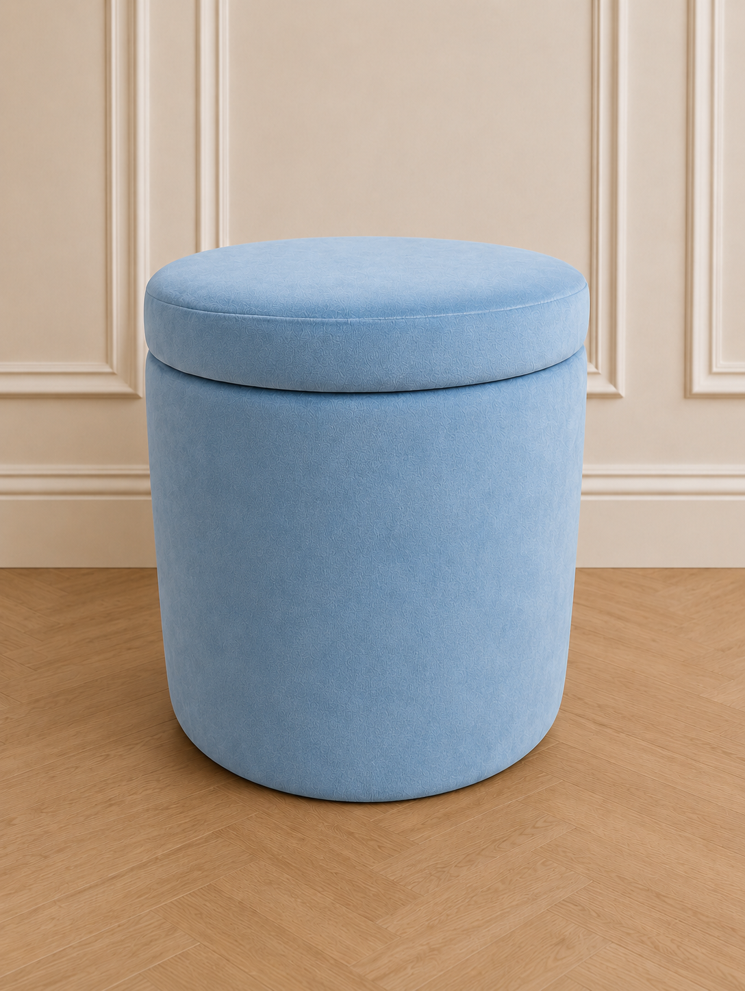 A light blue velvet cylindrical storage ottoman with a cushioned lid, set on a light wood herringbone floor against a beige paneled wall.