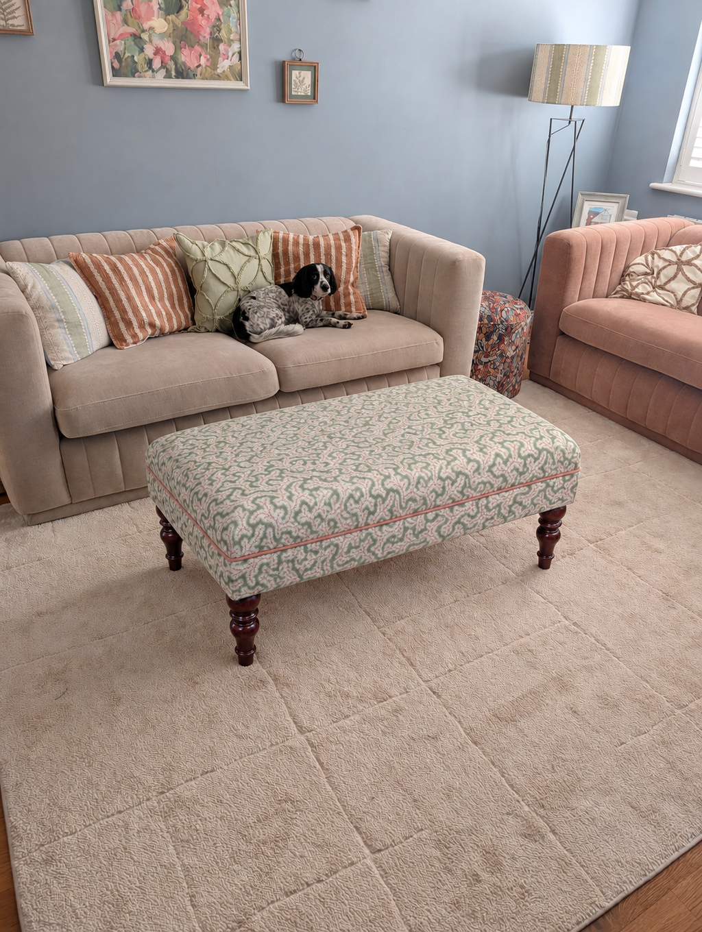 A large beige textured area rug in a styled living room, featuring a patterned upholstered ottoman, a beige sofa with striped pillows, and a pink armchair in the background.