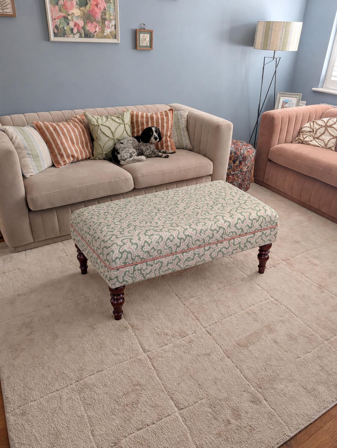 A large beige textured area rug in a styled living room, featuring a patterned upholstered ottoman, a beige sofa with striped pillows, and a pink armchair in the background.