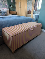 A rectangular upholstered storage bench with pink and beige vertical stripes, positioned at the foot of a bed in a bedroom setting.