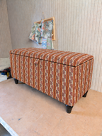 An upholstered storage bench with a striped pattern in rust orange, brown, and cream, standing on dark legs in an indoor room.