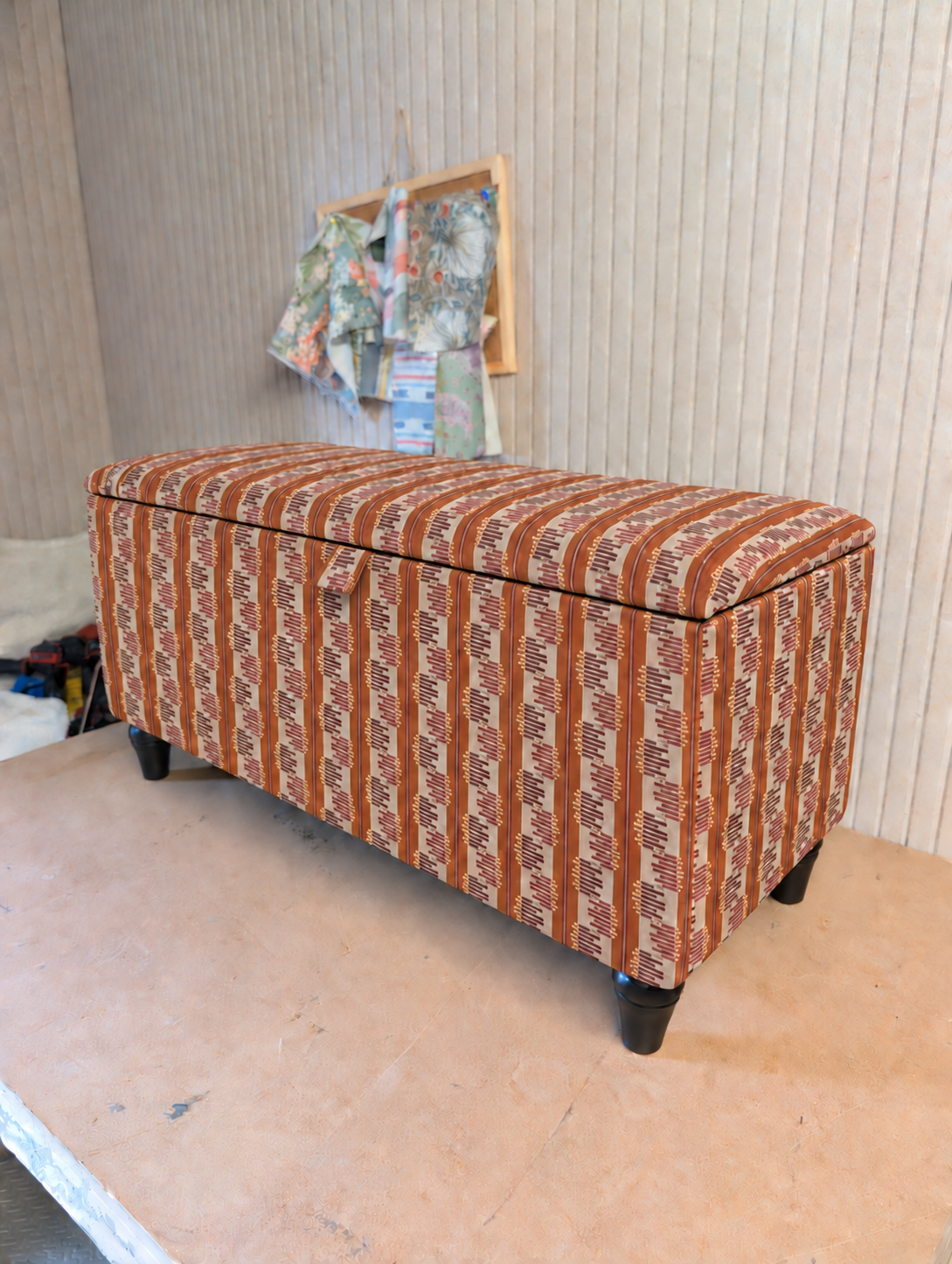 An upholstered storage bench with a striped pattern in rust orange, brown, and cream, standing on dark legs in an indoor room.