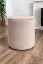 A round upholstered ottoman stool in a soft blush pink velvet fabric, set on a light brown wood floor in a modern room with white walls and a potted plant in the background.