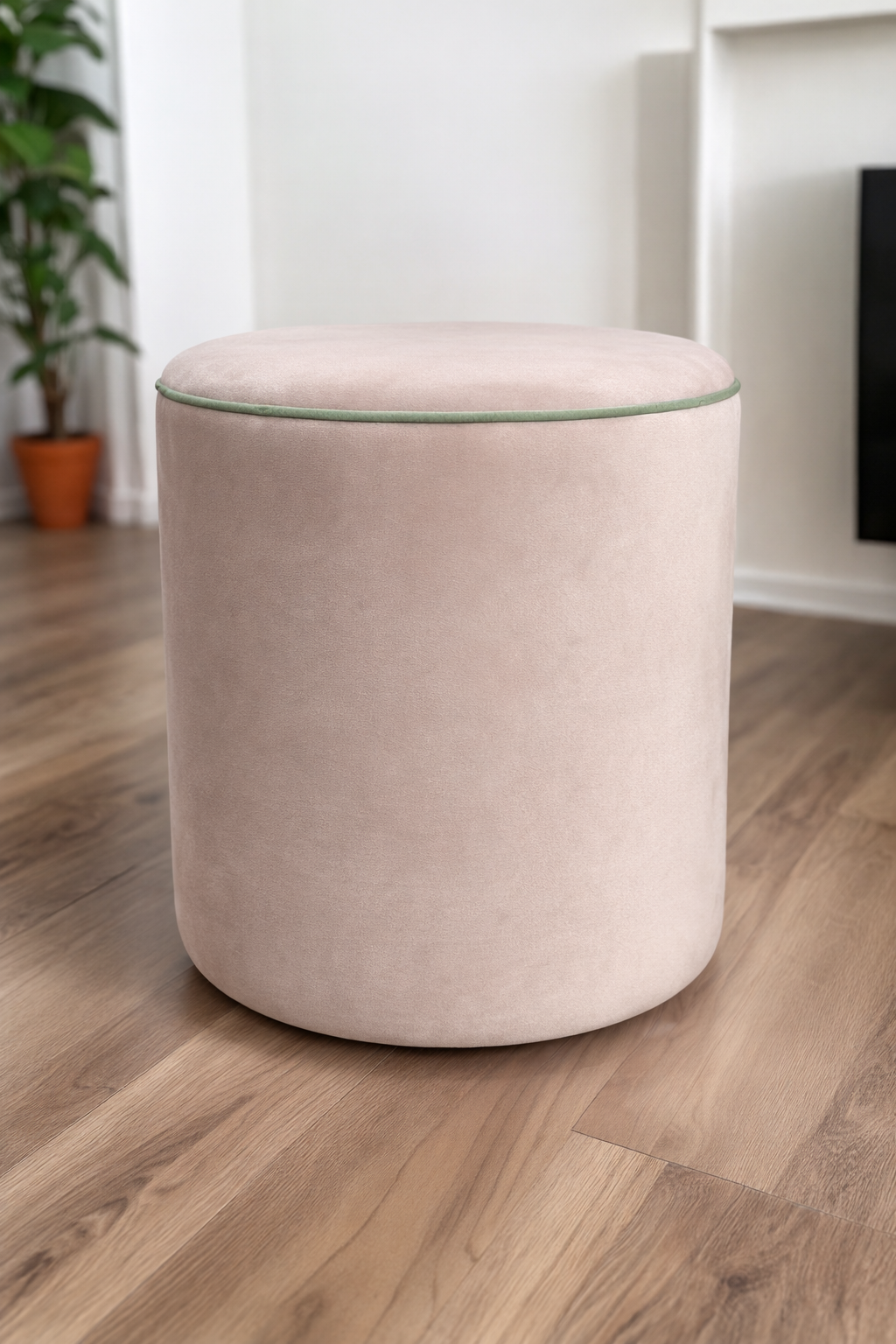 A round upholstered ottoman stool in a soft blush pink velvet fabric, set on a light brown wood floor in a modern room with white walls and a potted plant in the background.