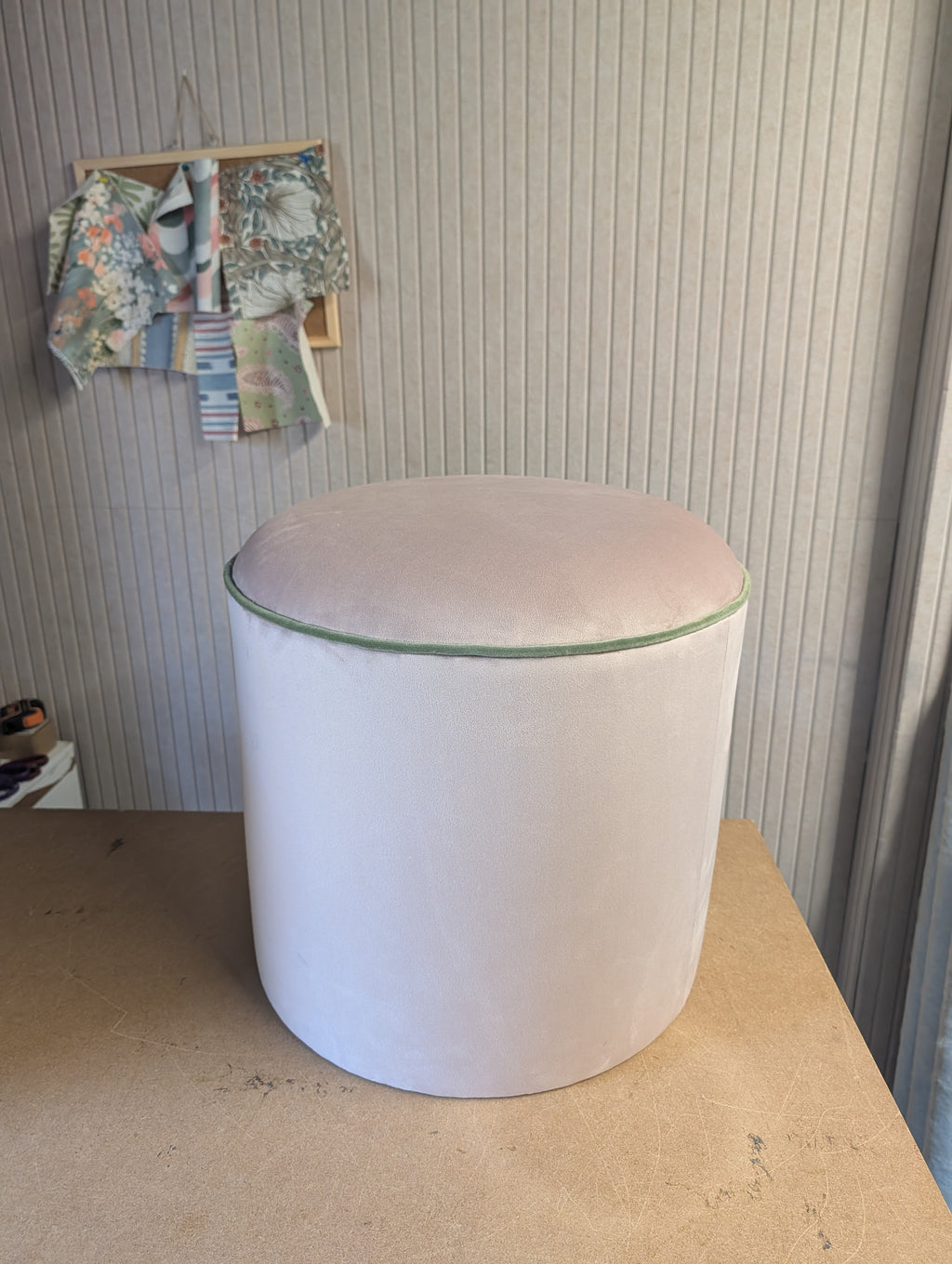 A cylindrical upholstered ottoman or stool with a pale blush pink velvet-like fabric and a thin green piping detail around the top edge, shown on a wooden table against a ribbed wall.