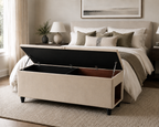 Beige upholstered storage bench with a dog or cat house hole in a bedroom with a bed and pillows in the background.