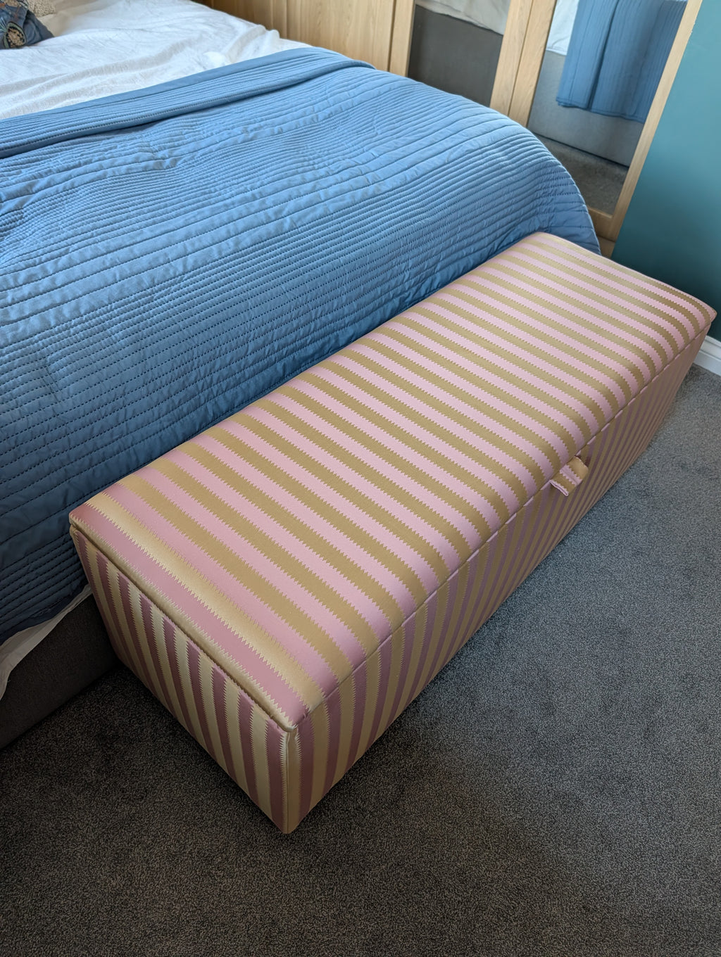 A rectangular upholstered storage bench with pink and yellow vertical stripes, positioned at the foot of a bed with a blue quilted cover in a bedroom setting.