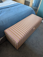 A rectangular upholstered storage bench with pink and yellow vertical stripes, positioned at the foot of a bed with a blue quilted cover in a bedroom setting.