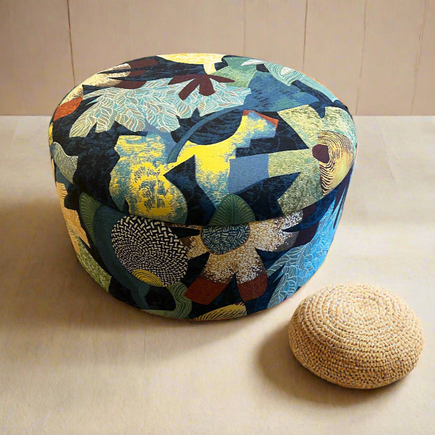 A round storage ottoman pouffe upholstered in a multicolored tropical fabric, with a beige woven footstool next to it.