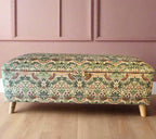 A rectangular storage ottoman upholstered in William Morris velvet with angled feet, featuring a floral pattern in a mix of gold, black, and green colors.