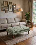 A styled living room featuring a beige fabric sofa and a green patterned upholstered ottoman on a large area rug, with a floor lamp, wall art, and a window in the background.