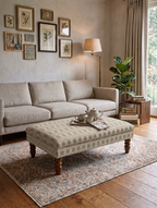 A styled living room featuring a light beige fabric sofa, a matching patterned upholstered coffee table with wooden legs, a large patterned area rug, a floor lamp, and framed botanical prints on the wall.
