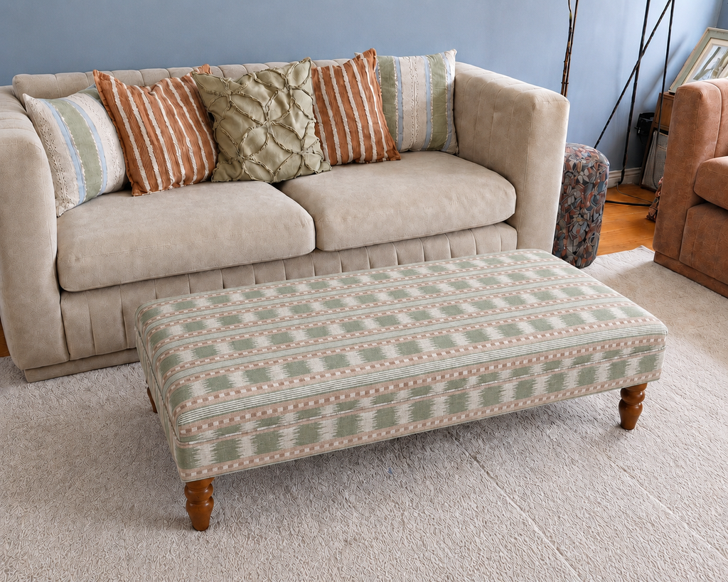 A patterned fabric ottoman with wooden legs placed in front of a beige sofa in a living room setting.