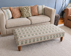 A patterned fabric ottoman with wooden legs placed in front of a beige sofa in a living room setting.