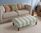 Handmade coffee table footstool upholstered in green, blue and cream Linwood Dimity fabric with turned wooden legs, styled in a neutral living room.
