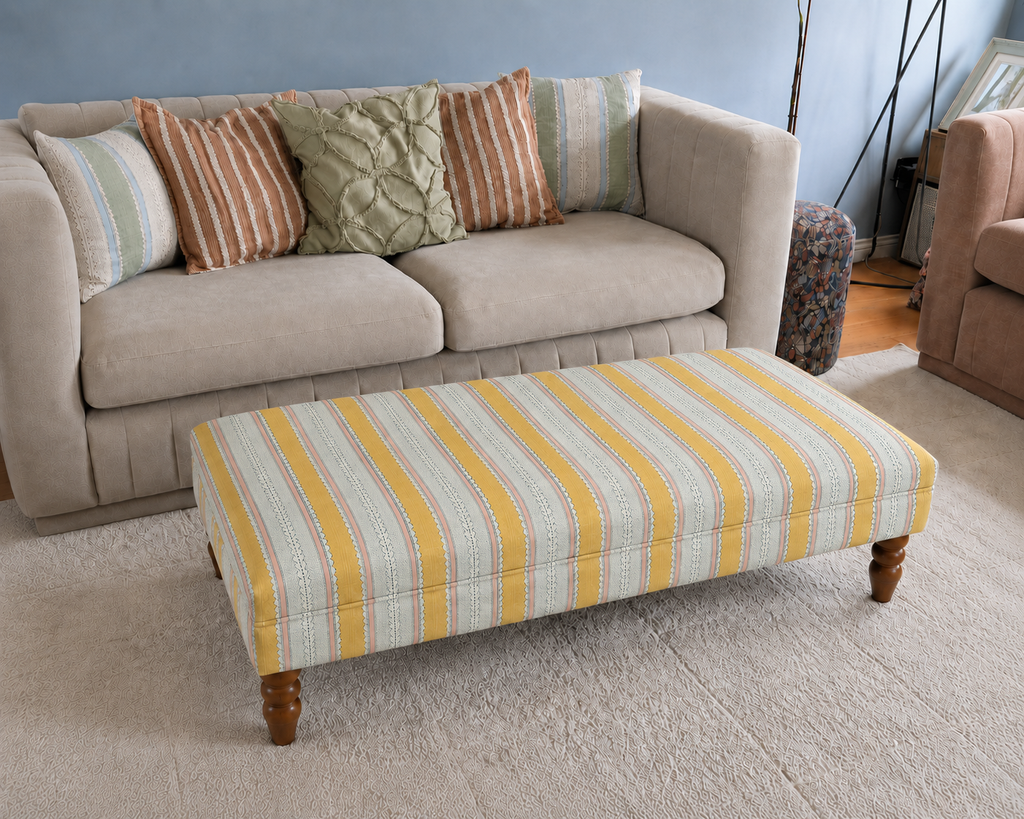 Handmade upholstered footstool in yellow, blue and cream Linwood Dimity striped fabric, styled in front of a neutral sofa.