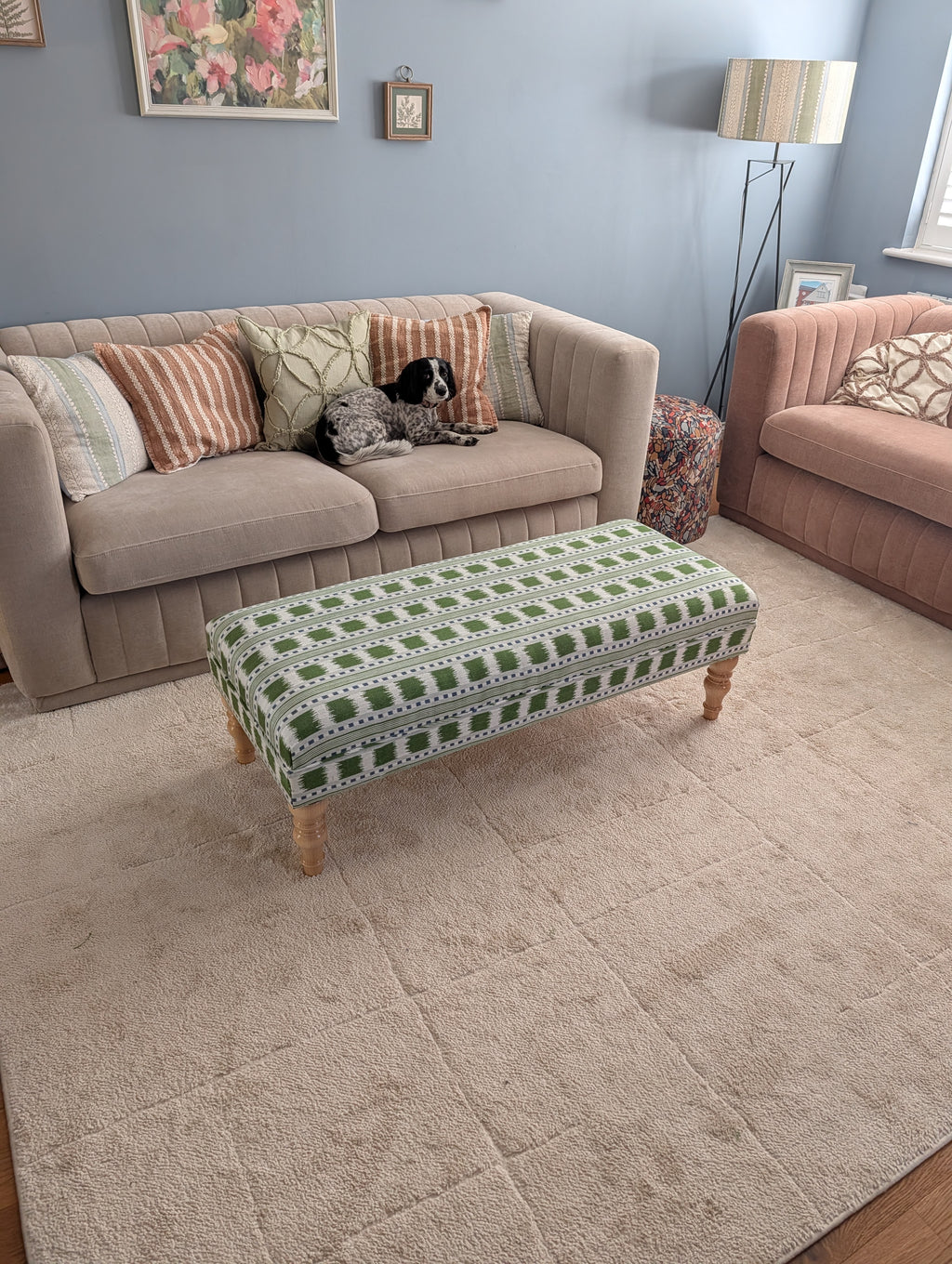 A styled living room scene featuring a large beige textured area rug under a patterned green and white upholstered ottoman bench. In the background, there is a beige sofa with striped and floral pillows, a pink armchair, and a blue-gray wall.