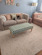 A styled living room scene featuring a large beige textured area rug under a patterned green and white upholstered ottoman bench. In the background, there is a beige sofa with striped and floral pillows, a pink armchair, and a blue-gray wall.