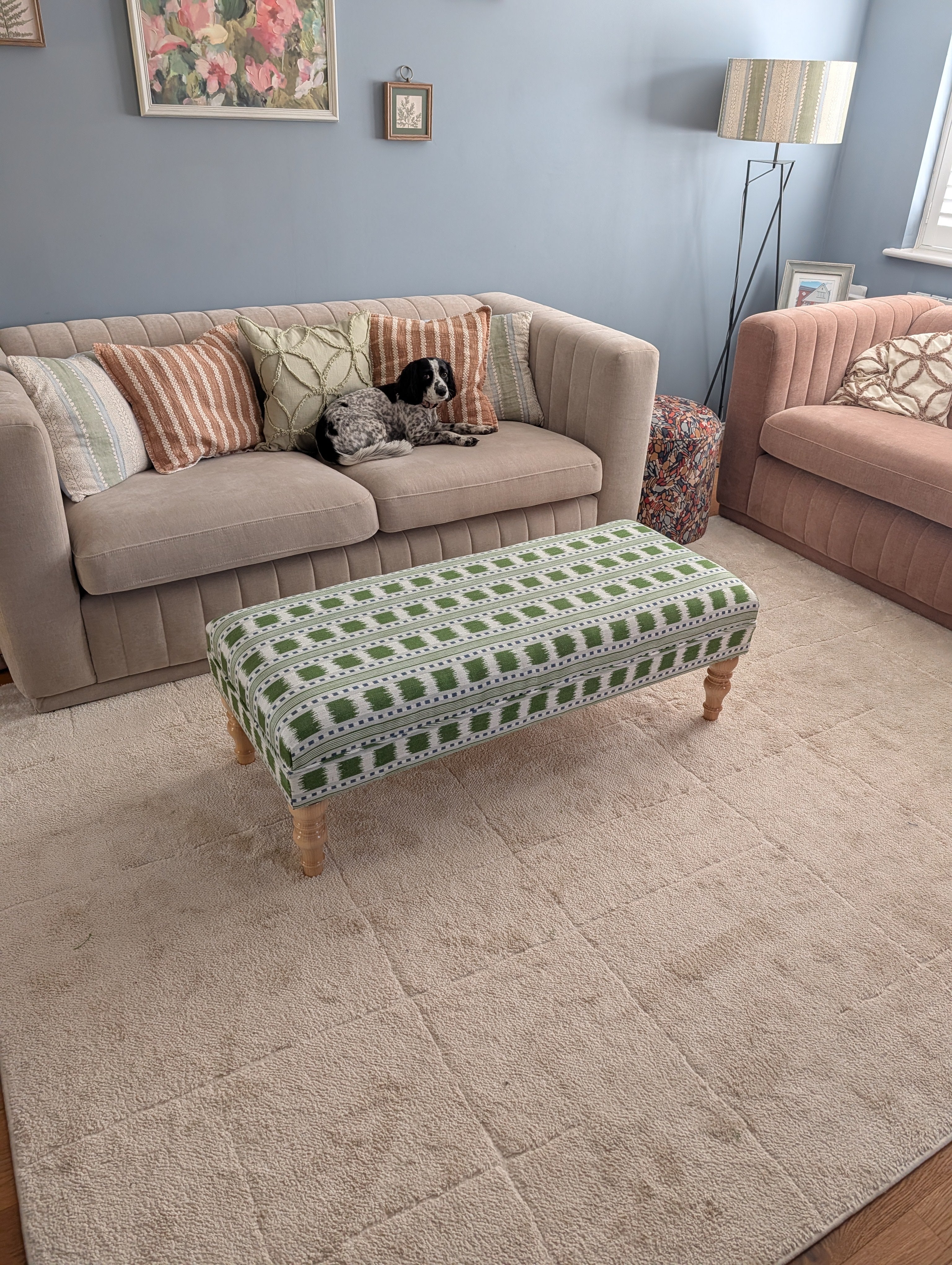 A styled living room scene featuring a large beige textured area rug under a patterned green and white upholstered ottoman bench. In the background, there is a beige sofa with striped and floral pillows, a pink armchair, and a blue-gray wall.