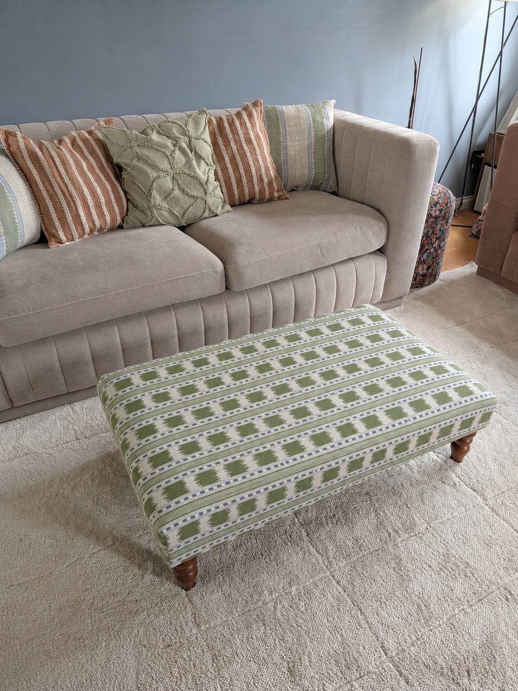 A rectangular upholstered ottoman with a green and white geometric pattern and small wooden legs, placed in front of a beige sofa on a light rug.