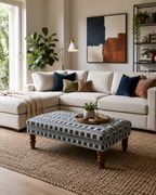 A styled living room featuring a cream sectional sofa with navy, olive, and brown accent pillows, a matching patterned ottoman coffee table, a large woven area rug, and a beige wall with abstract wall art.