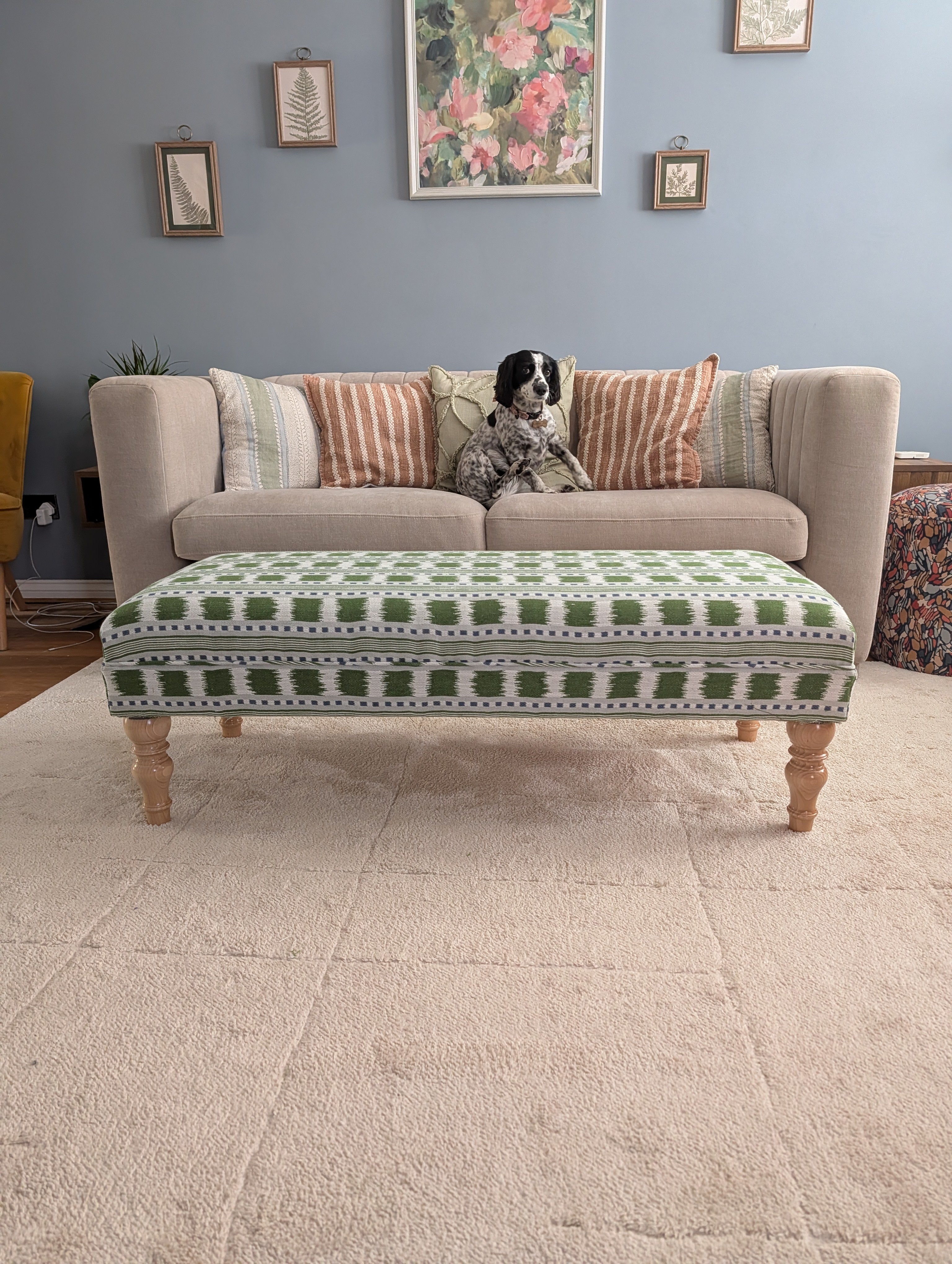 A green and white patterned upholstered ottoman with wooden legs sits in front of a beige sofa in a living room. A black and white dog is sitting on the sofa, and the room has a blue-gray wall, framed art, and a cream-colored rug.